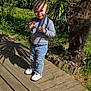 child, toddler, outdoor, sunlight, shadows, wooden_deck, bicycle, fence, greenery, plants, grass, jeans, sneakers, gray_sweater, smile, playful, person, nature, daylight, garden