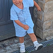 Aléssio participe au concours pour gagner de l'argent avec cette photo : child, boy, smiling, blue_clothing, shorts, button_shirt, blond_hair, white_sneakers, socks, outdoor, stone_wall, door, playful, standing, pavement, curb, grin, young, casual, happy