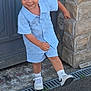 child, boy, smiling, blue_clothing, shorts, button_shirt, blond_hair, white_sneakers, socks, outdoor, stone_wall, door, playful, standing, pavement, curb, grin, young, casual, happy