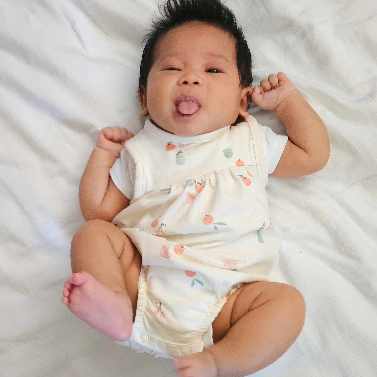 Kiri is registered to the contest to win money with this photo: adorable, arms, baby, child, cute, expression, foot, fruit_pattern, hair, hand, indoors, infant, legs, lying_down, playful, romper, skin, tongue_out, white_bed, winking