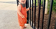 Benny joined the competition — help win amazing prizes! baby, child, toddler, smiling, outdoor, portrait, fence, garden, plants, park, walkway, orange_romper, striped_clothing, barefoot, infant, standing, happy, daylight, wheelchair, urban_landscape