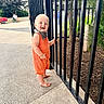 baby, child, toddler, smiling, outdoor, portrait, fence, garden, plants, park, walkway, orange_romper, striped_clothing, barefoot, infant, standing, happy, daylight, wheelchair, urban_landscape