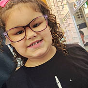 Ezra is registered to the contest to win money with this photo: child, girl, curly_hair, pink_bow, glasses, eyewear_store, smile, black_shirt, skeleton_hand, portrait, indoor, fashion, cute, happy, person, eyeglasses, apparel, closeup, retail, shopping