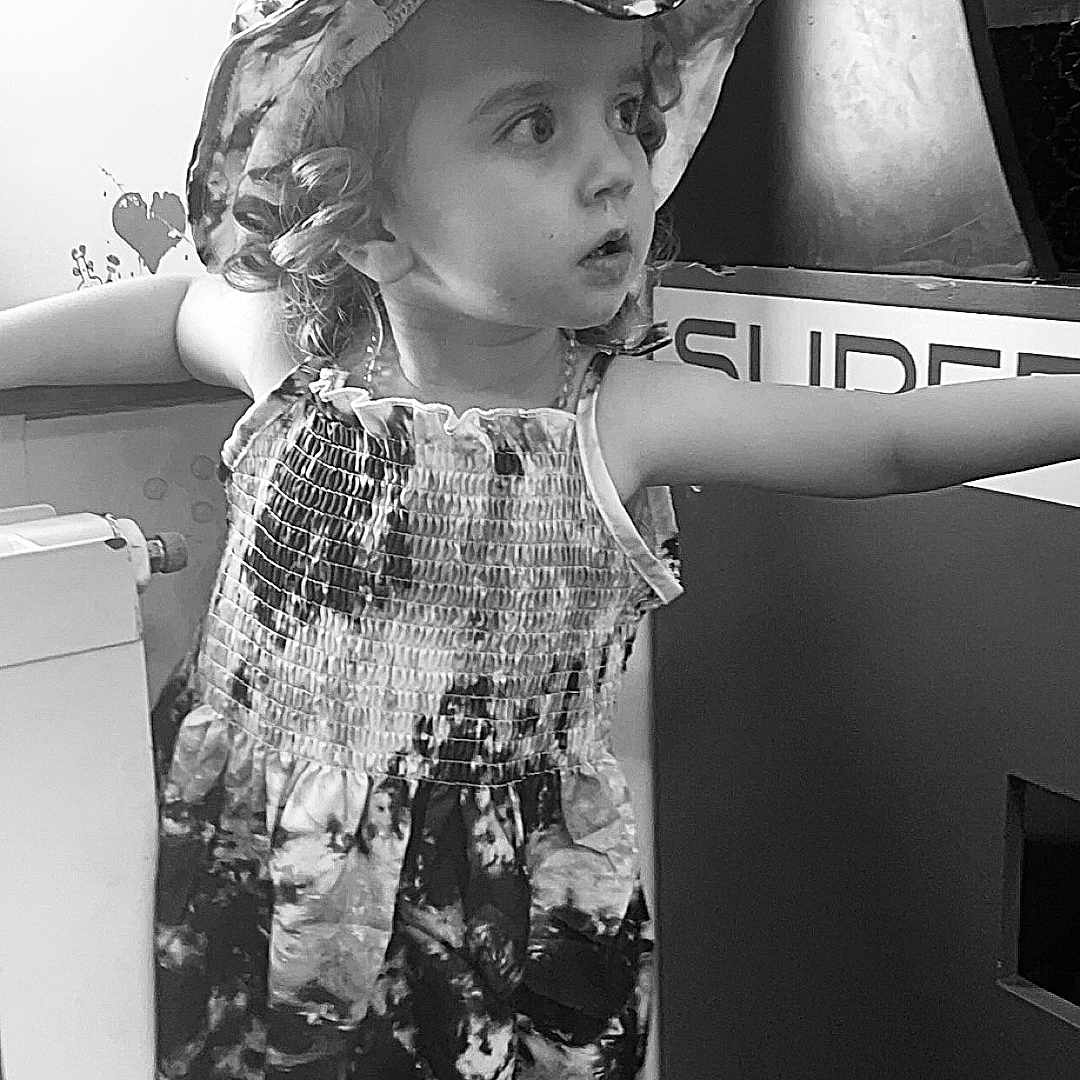 Ylana participe au concours pour gagner de l'argent avec cette photo : arms_outstretched, black_and_white, child, curiosity, curly_hair, dress, expression, floor, girl, hat, indoors, looking_away, monochrome, patterned_clothing, person, portrait, radiator, standing, wall, young_child