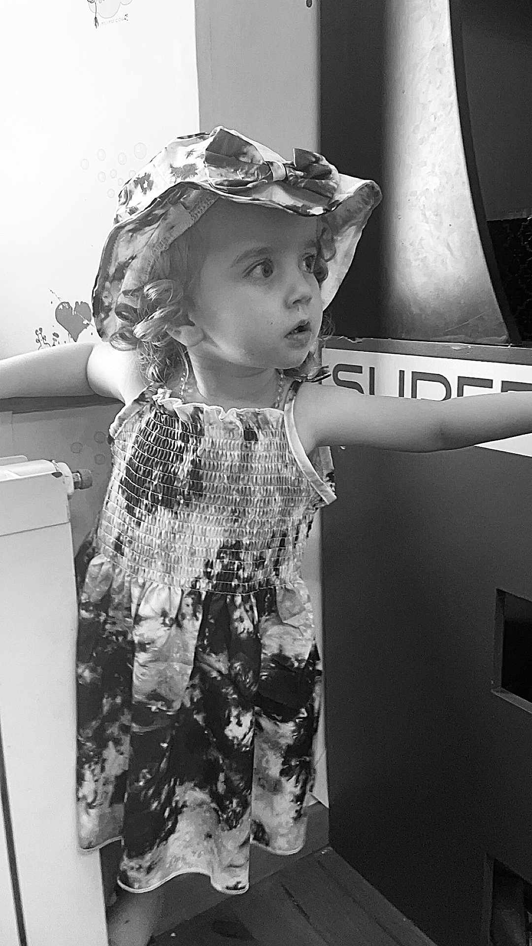 Ylana participe au concours pour gagner de l'argent avec cette photo : child, girl, hat, dress, curly_hair, black_and_white, indoors, expression, portrait, standing, floor, wall, radiator, patterned_clothing, person, young_child, looking_away, arms_outstretched, curiosity, monochrome