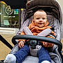 baby, stroller, child, smile, orange_jacket, navy_pants, white_socks, outdoor, cafe, window, seat_belt, happy, infant, person, portrait, cozy, casual_clothing, urban, daytime, cute