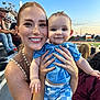 woman, baby, smiling, outdoor, stadium, sunset, crowd, clothing, person, happy, portrait, child, holding, blue_shirt, checkered_shorts, nature, event, people, seated, family
