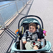 Yazid is registered to the contest to win money with this photo: baby, child, stroller, smiling, outdoor, waterfront, cityscape, sidewalk, railing, sunlight, shadows, portrait, infant, seated, plaid_shirt, green_pants, toy, urban, skyline, happy