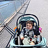 baby, child, stroller, smiling, outdoor, waterfront, cityscape, sidewalk, railing, sunlight, shadows, portrait, infant, seated, plaid_shirt, green_pants, toy, urban, skyline, happy