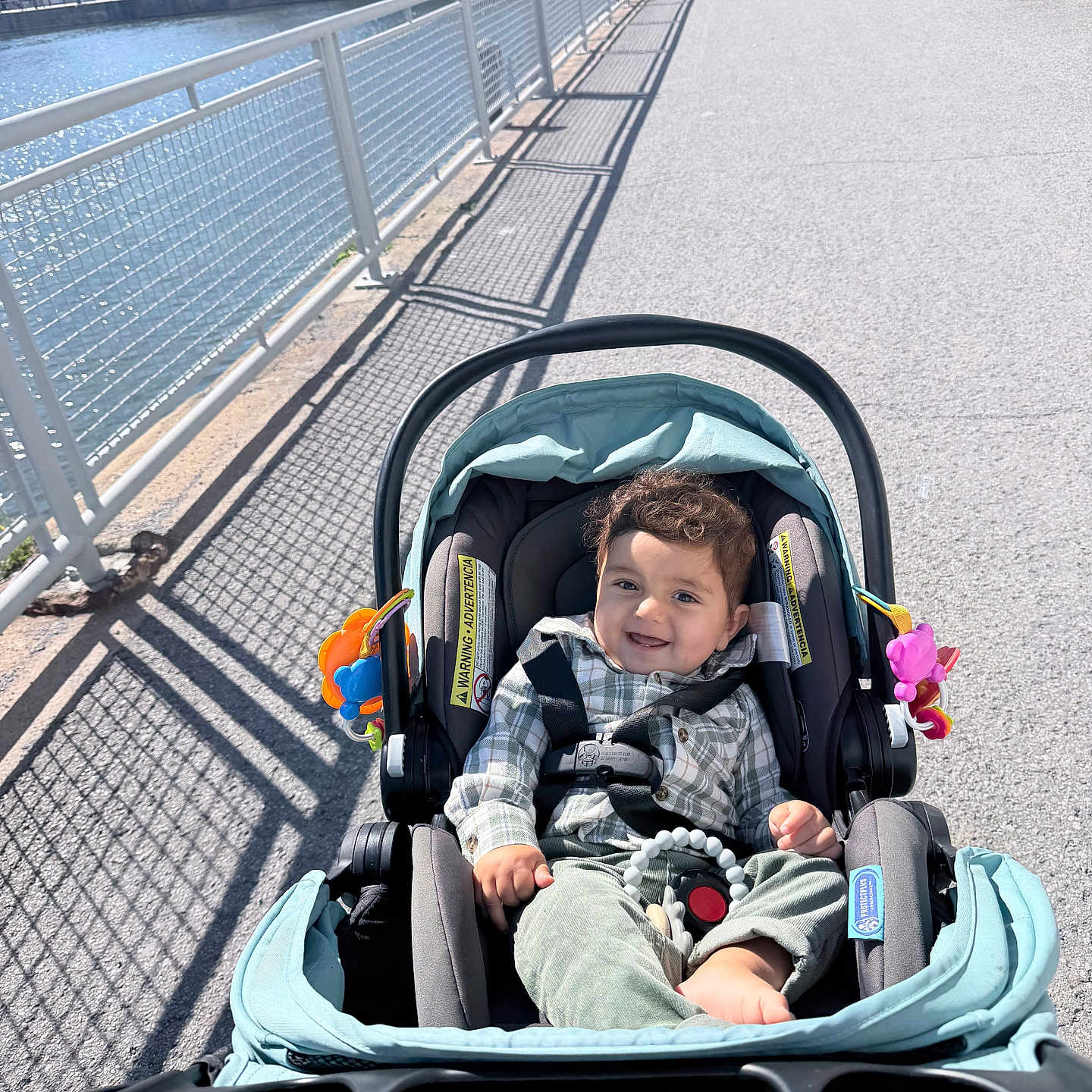 Yazid is registered to the contest to win money with this photo: baby, child, stroller, smiling, outdoor, waterfront, cityscape, sidewalk, railing, sunlight, shadows, portrait, infant, seated, plaid_shirt, green_pants, toy, urban, skyline, happy