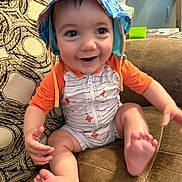 Carson joined the competition — help win amazing prizes! baby, child, smiling, hat, onesie, couch, indoor, happy, sitting, feet, hand, face, patterned_pillow, clothing, infant, cute, expression, living_room, furniture, colorful