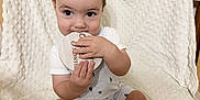 Carson is registered to the contest to win money with this photo: baby, child, infant, sitting, blanket, wooden_disc, teething, cute, indoors, person, clothing, shorts, white_shirt, face, hand, foot, texture, light, skin, expression