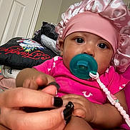 Keauni is registered to the contest to win money with this photo: baby, infant, pacifier, pink_bonnet, pink_clothing, hand, adult_hand, nail_polish, indoor, door, pillow, child, closeup, face, skin, cute, holding, comfort, young, person