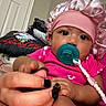 baby, infant, pacifier, pink_bonnet, pink_clothing, hand, adult_hand, nail_polish, indoor, door, pillow, child, closeup, face, skin, cute, holding, comfort, young, person