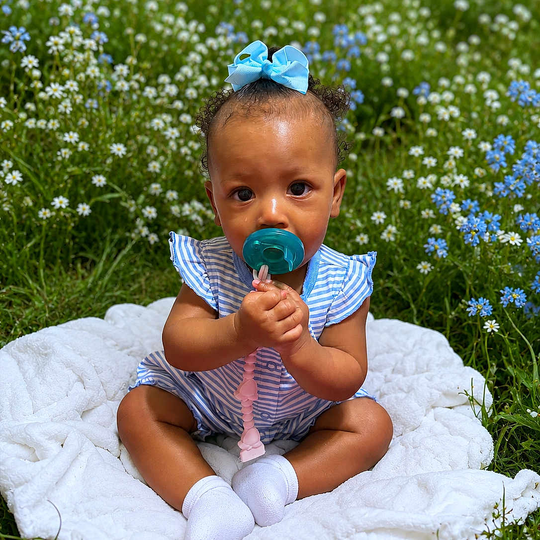 Keauni is registered to the contest to win money with this photo: baby, child, pacifier, blue_bow, striped_clothing, white_blanket, wildflowers, grass, outdoor, cute, sitting, infant, nature, greenery, blue_flowers, white_flowers, curly_hair, sock, summer, portrait