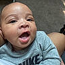 Princeston is registered to the contest to win money with this photo: baby, infant, child, portrait, close_up, face, smiling, open_mouth, wide_eyed, gray_shirt, clothing, text_on_clothing, adult_holding, indoor, home, skin, head, hands, sitting, cute