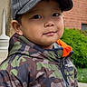 child, boy, cap, jacket, camo, camouflage, outdoor, smile, face, person, greenery, brick_wall, urban, young, portrait, casual, hooded_jacket, nature, curious, cute