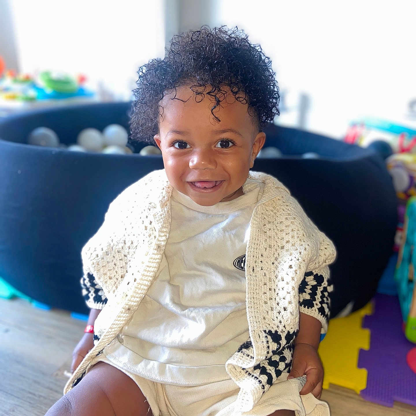 Neals a rejoint le concours — aidez-le/la à gagner de superbes lots ! ball_pit, bright_light, casual_clothing, child, curly_hair, cute, happy, home, indoor, knitted_cardigan, person, play_area, portrait, sitting, smiling, toddler, toy, white_clothing, wooden_floor, young_child