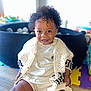 toddler, child, smiling, curly_hair, knitted_cardigan, white_clothing, indoor, wooden_floor, play_area, ball_pit, happy, portrait, cute, sitting, young_child, person, bright_light, home, toy, casual_clothing