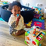 toddler, child, curly_hair, knitted_sweater, toy, chewing, wooden_floor, ball_pit, colorful_play_mats, activity_cube, indoor, playtime, smiling, happy, footwear, shoes, bright_colors, person, candid, cute