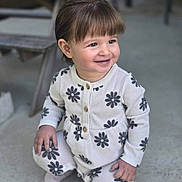 Emy a rejoint le concours — aidez-le/la à gagner de superbes lots ! toddler, child, smiling, kneeling, outdoor, concrete_floor, white_clothing, flower_pattern, long_sleeves, pants, brown_hair, cute, happy, person, portrait, casual, daylight, small_hands, buttoned_top, cozy_outfit