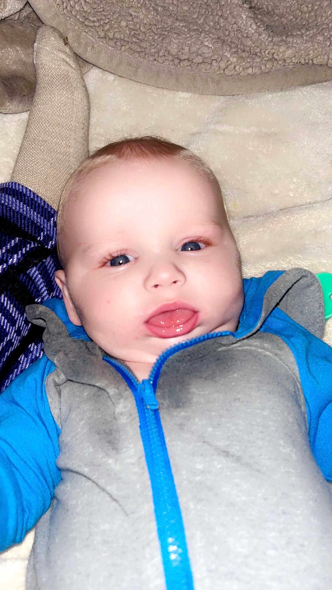 Isaiah joined the competition — help win amazing prizes! baby, infant, blue_clothing, gray_clothing, zipper, face, tongue, blanket, soft_texture, lying_down, close_up, child, cute, indoors, happy, person, toddler, smiling, skin, head