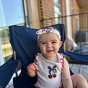 Margo is registered to the contest to win money with this photo: baby, child, smiling, headband, cherry, white_top, navy_chair, porch, wooden_floor, outdoor, sunlight, happy, cute, person, sitting, infant, summer, fashion, playful, closeup