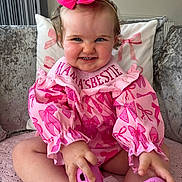 Darcey is registered to the contest to win money with this photo: baby, child, pink, bow, smile, couch, cushion, clothing, footwear, indoor, person, cute, happy, toddler, hair, sitting, hand, face, playful, home