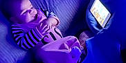 Octavia is registered to the contest to win money with this photo: baby, infant, tablet, screen, blanket, pillow, indoor, blue_light, striped_clothing, sleepwear, child, technology, couch, nighttime, relaxing, portrait, smiling, soft_lighting, resting, cute