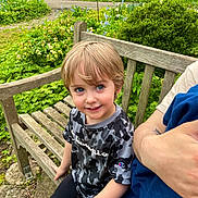 Zyah is registered to the contest to win money with this photo: child, bench, garden, flowers, greenery, smiling, blonde_hair, blue_eyes, camouflage_shirt, champion_brand, outdoor, person, wood, nature, casual_clothing, happy, portrait, daylight, adult_arm, candid