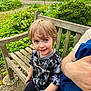 child, bench, garden, flowers, greenery, smiling, blonde_hair, blue_eyes, camouflage_shirt, champion_brand, outdoor, person, wood, nature, casual_clothing, happy, portrait, daylight, adult_arm, candid