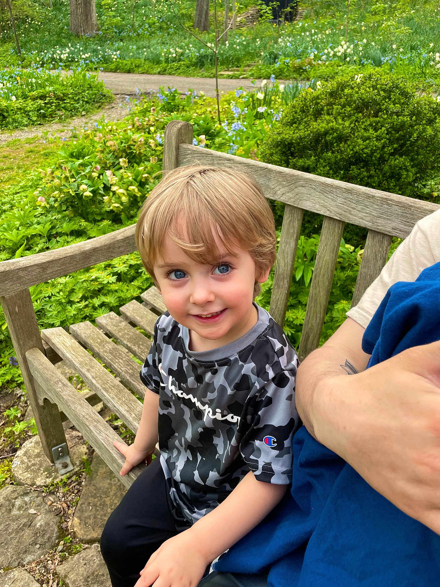 Zyah is registered to the contest to win money with this photo: child, bench, garden, flowers, greenery, smiling, blonde_hair, blue_eyes, camouflage_shirt, champion_brand, outdoor, person, wood, nature, casual_clothing, happy, portrait, daylight, adult_arm, candid