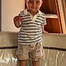 toddler, child, person, standing, striped_shirt, polo_shirt, denim_shorts, ripped_shorts, sneakers, white_shoes, couch, indoor, wood_paneling, smiling, short_hair, cute, fashion, casual_clothing, young_child, confident
