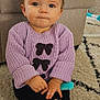 baby, child, sitting, purple_sweater, yellow_bow, pony_tail, blue_eyes, carpet, toy_comb, socks, bear_face, indoor, furniture, curious_expression, cozy_clothing, hands, floor, playful, cute, person