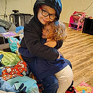 Raina joined the competition — help win amazing prizes! child, children, hug, smile, glasses, hoodie, curly_hair, indoor, wooden_floor, blanket, toy, sneakers, sweater, pants, couch, person, warm_clothing, happy, home, playroom