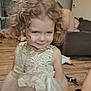 child, girl, curly_hair, dress, smile, face, indoor, living_room, toy, floor, couch, person, playful, young_child, portrait, home, wood_floor, furniture, expression, cute
