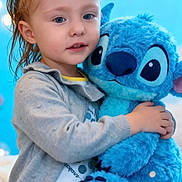 Enola a rejoint le concours — aidez-le/la à gagner de superbes lots ! child, toddler, plush_toy, blue, yellow_flower, hair_clip, hugging, soft_toy, polka_dot, cardigan, indoor, cute, smiling, portrait, cozy, bedroom, playful, happy, toy, blue_background