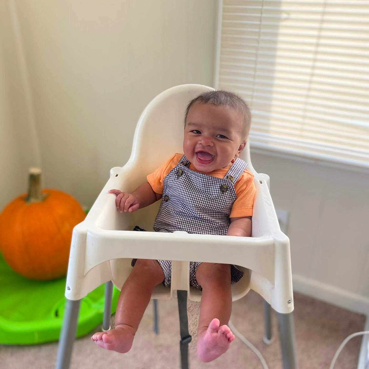 Zayn is registered to the contest to win money with this photo: baby, carpet, checkered_overalls, child, cute, feet, furniture, happy, high_chair, home, indoor, infant, light, orange_shirt, person, portrait, pumpkin, seat, smiling, window_blinds