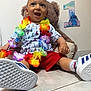 toddler, child, smiling, sitting, flower_lei, red_shorts, white_sneakers, teddy_bear, birthday, decorations, floor, tile_floor, wall, stitch, celebration, happy, indoors, person, cute, colorful