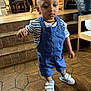 toddler, child, blue_overalls, white_sneakers, striped_shirt, indoor, tile_floor, stairs, wooden_furniture, person_walking, necklace, baby, footwear, floor, person, home, casual_clothing, young_child, walking, expression