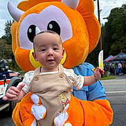Dylan is registered to the contest to win money with this photo: baby, child, infant, mascot, dinosaur, triceratops, costume, outdoor, festival, portrait, face, smiling, sitting, overalls, bib, toy, orange, blue, street, daylight