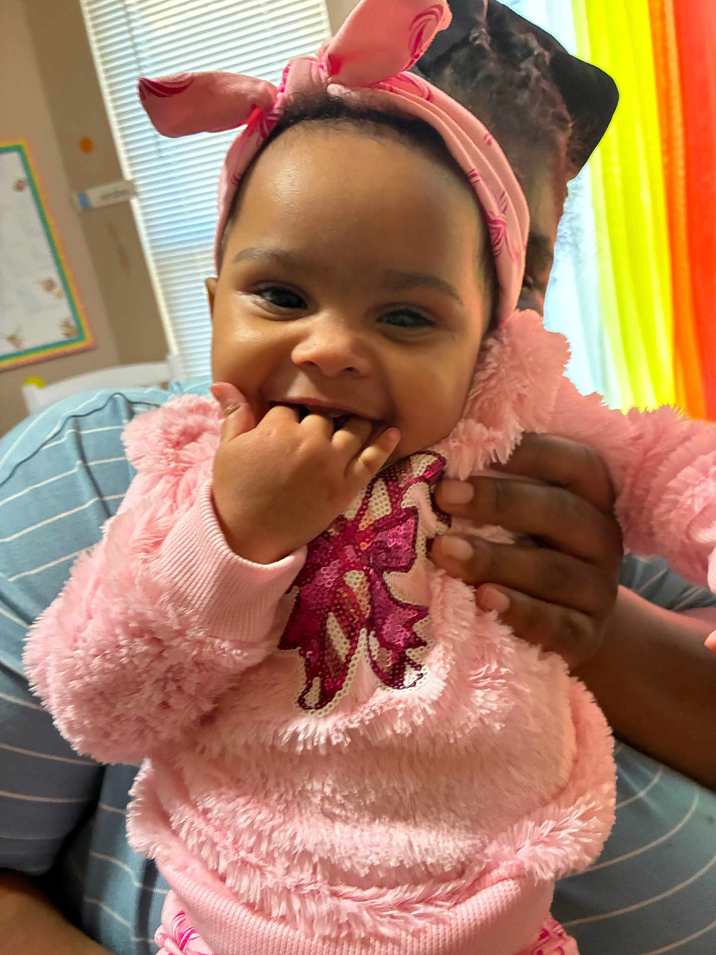 Maverick joined the competition — help win amazing prizes! baby, child, pink_clothing, headband, smiling, hand, adult_hand, indoor, window, curtains, striped_shirt, person, happy, cute, toddler, playful, seated, face, fingers_in_mouth, cozy