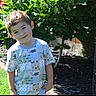 child, boy, smiling, garden, plants, greenery, sunlight, outdoor, grass, flower_bed, tree, shirt, patterned_clothing, happy, young, nature, daylight, casual, portrait, playful