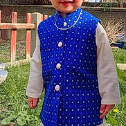 Kabir is registered to the contest to win money with this photo: blue_vest, child, daylight, face, fence, garden, grass, happy, nature, necklace, outdoor, person, portrait, smiling, standing, sunlight, traditional_clothing, tree, white_pants, young