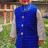 child, smiling, outdoor, garden, grass, fence, tree, traditional_clothing, blue_vest, white_pants, necklace, face, person, sunlight, nature, happy, standing, portrait, young, daylight
