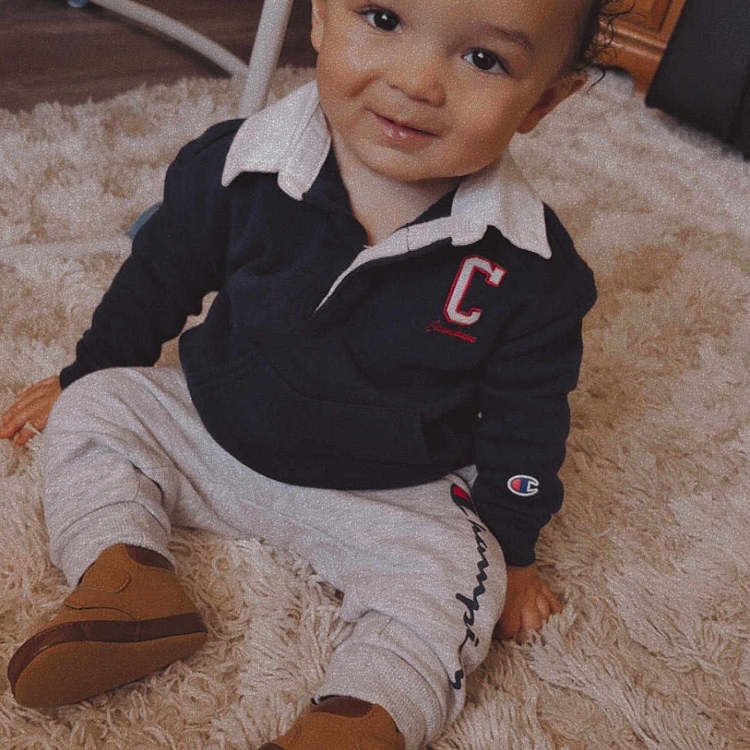 Isaiah is registered to the contest to win money with this photo: baby, carpet, casual, child, clothing, curly_hair, cute, furniture, happy, indoors, pants, person, portrait, rug, shoes, sitting, smiling, sweatshirt, toddler, wooden_drawer