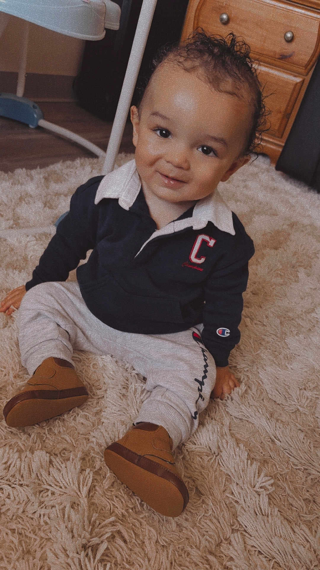 Isaiah is registered to the contest to win money with this photo: toddler, child, smiling, curly_hair, sitting, rug, indoors, clothing, sweatshirt, pants, shoes, carpet, furniture, wooden_drawer, happy, cute, portrait, baby, person, casual