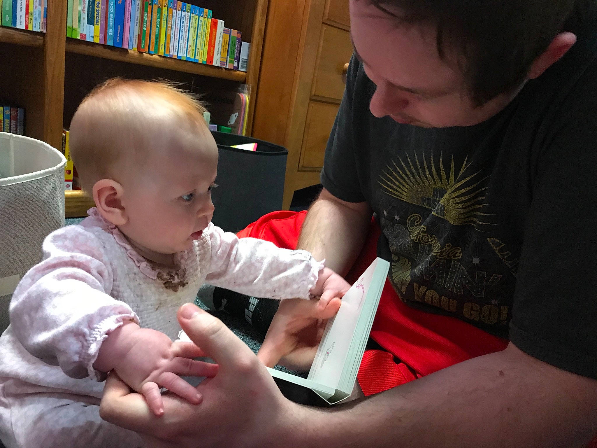 Madeline is registered to the contest to win money with this photo: arm, baby, baby_toddler_clothing, bookcase, carmine, child, comfort, fun, gesture, hair, hand, lap, person, play, publication, sharing, shelf, shelving, sitting, t_shirt