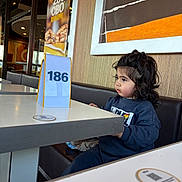 Izak participe au concours pour gagner de l'argent avec cette photo : child, toddler, restaurant, table, table_number, curly_hair, indoor, seated, casual_clothing, booth, wall_art, wood_paneling, curious, portrait, waiting, young_child, face, hair, furniture, person