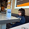 child, toddler, restaurant, table, table_number, curly_hair, indoor, seated, casual_clothing, booth, wall_art, wood_paneling, curious, portrait, waiting, young_child, face, hair, furniture, person